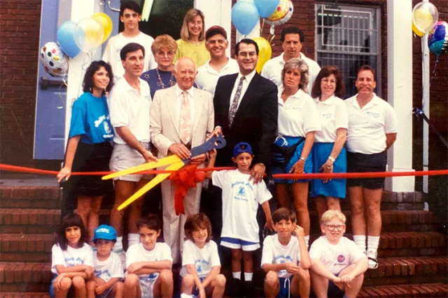 A group of adults and children pose for a photo at a ribbon-cutting ceremony outside a brick building, with balloons and oversized scissors displayed.