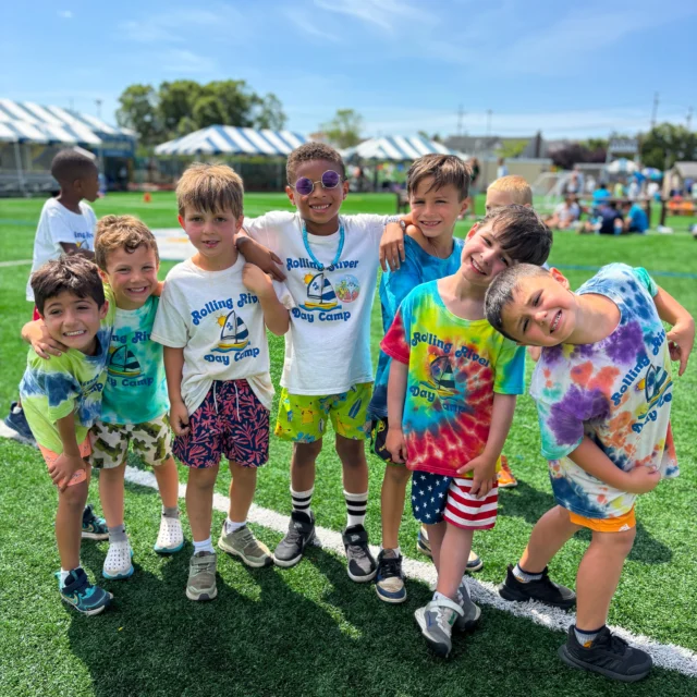 A group of young boys wearing colorful shirts and shorts stand together and smile on a grassy field at a summer day camp.