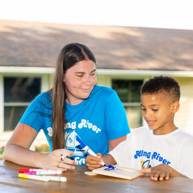 A woman and a young boy sit at an outdoor table, drawing on paper with markers. Both wear "Rolling River" shirts. A building is visible in the background.