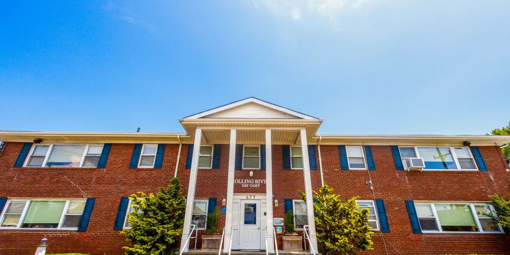 A two-story red brick building with white columns, blue shutters, and a sign above the entrance that reads "Rolling Ridge on 3rd." Steps lead up to the main door, flanked by shrubs.