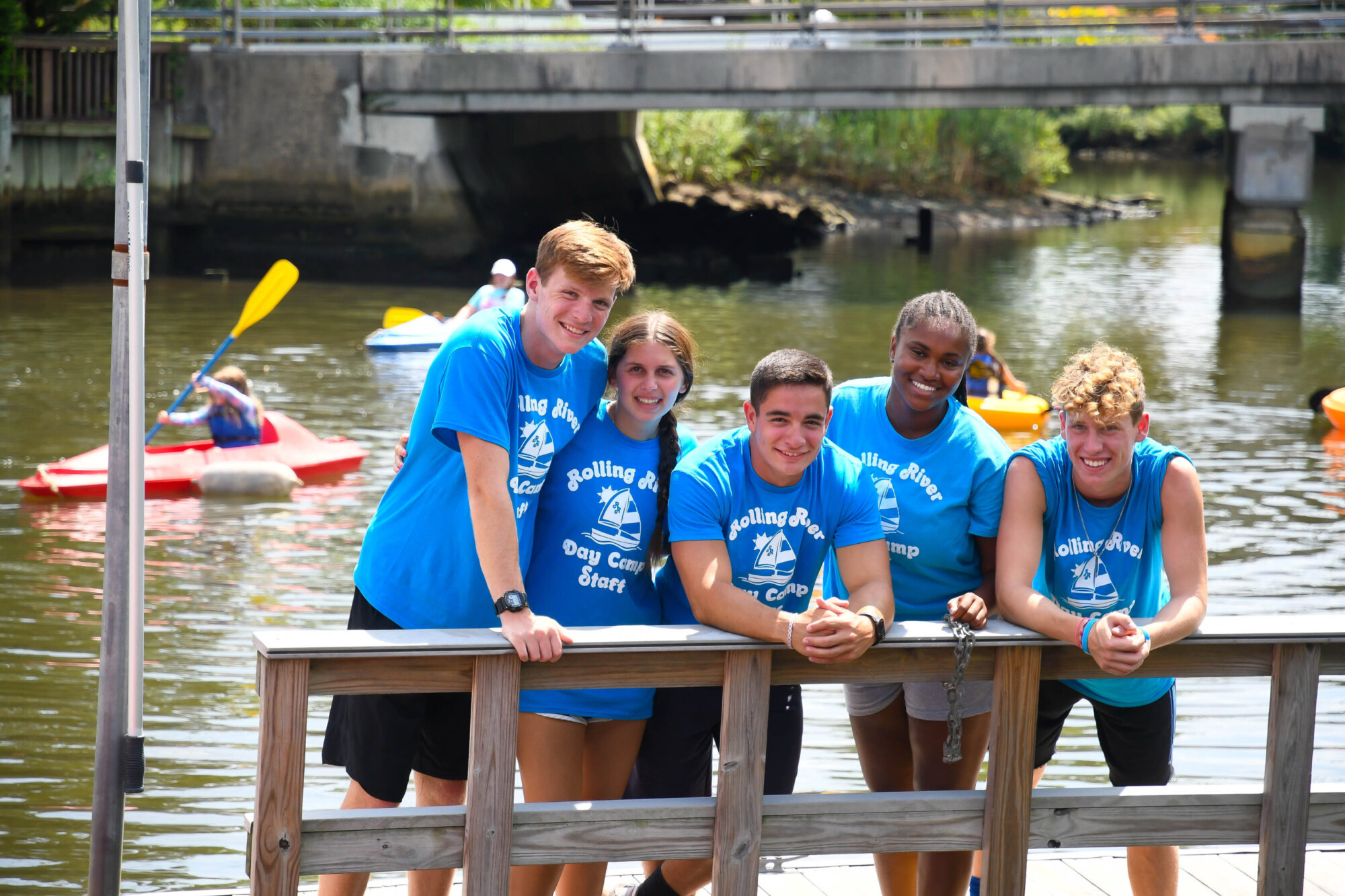 Five teenagers wearing matching blue camp shirts lean against a wooden railing by the water, with kayakers paddling in the background.