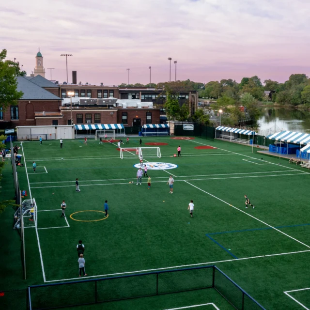 Aerial view of a sports complex with multiple soccer fields, tents along the side, and people playing soccer under a pink and blue sky.