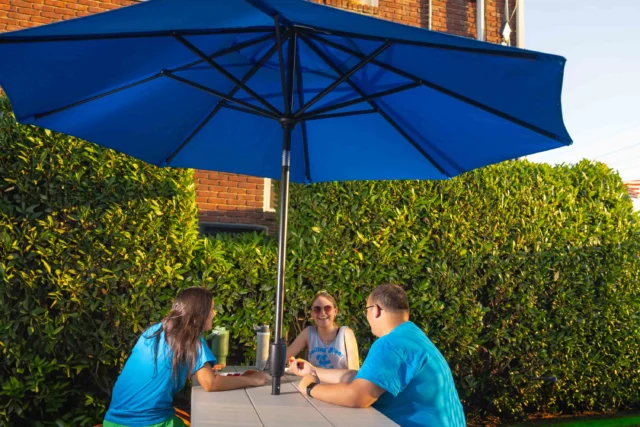 Three people sit at an outdoor table under a large blue umbrella, talking and enjoying the sunny weather. Bushes and a brick building are in the background.
