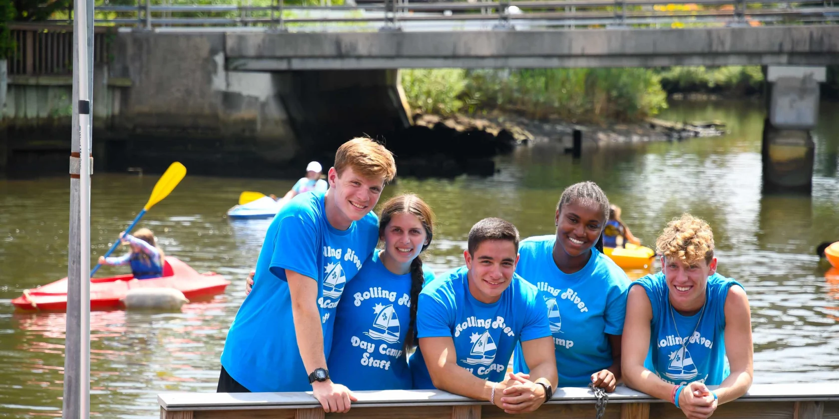 Five young people in matching blue camp t-shirts stand and lean on a wooden railing by the water, with kayakers paddling in the background.