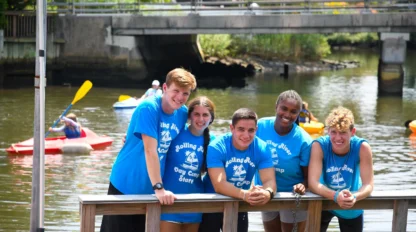 Five young people in matching blue camp t-shirts stand and lean on a wooden railing by the water, with kayakers paddling in the background.