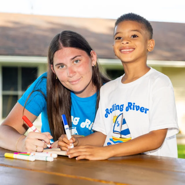 A woman and a boy sit at a wooden table outside, smiling and drawing with markers in front of a house.