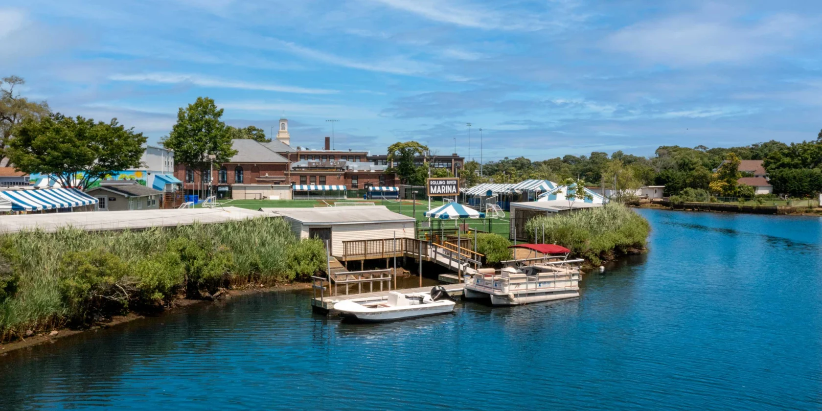 A small marina with docked boats along a calm river, surrounded by green vegetation and buildings with striped awnings under a blue sky.