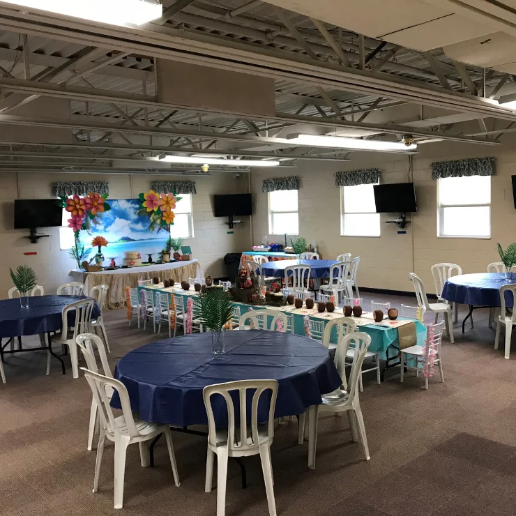 A decorated event room with round tables covered in blue tablecloths and white chairs, featuring a tropical-themed setup and colorful wall decorations.