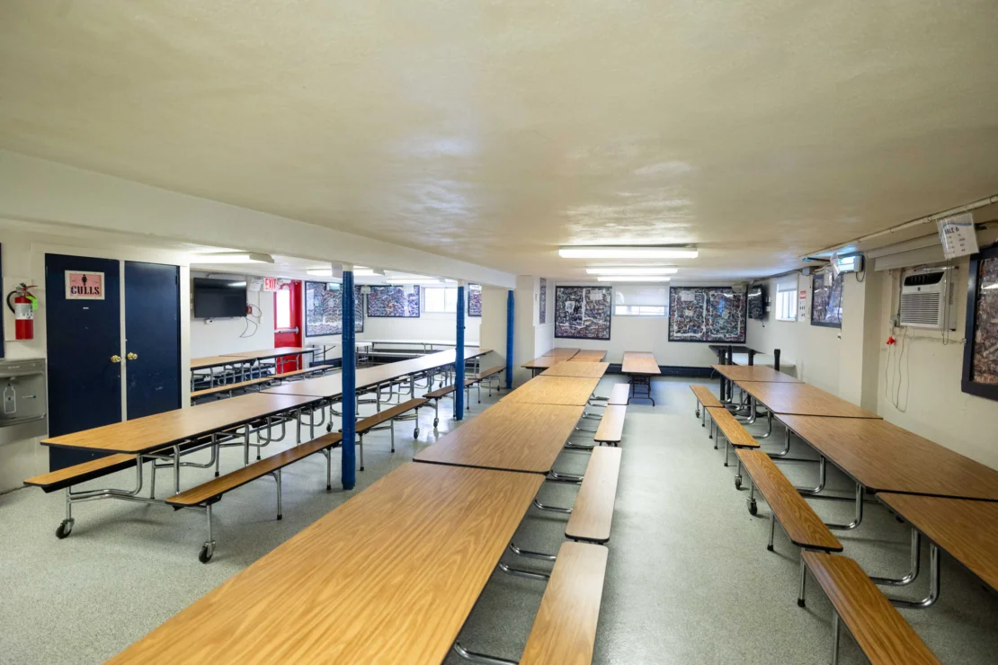 A school cafeteria with long wooden tables and attached benches, empty of people, fluorescent lighting, and posters on the walls.