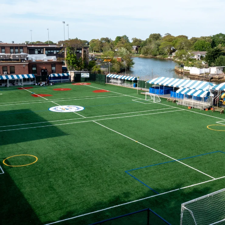 A green artificial turf soccer field with white lines, blue and white striped tents on the side, and a building and waterway in the background.