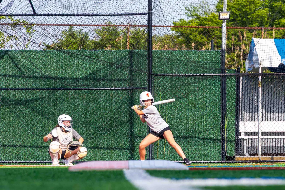 A softball batter prepares to swing at a pitch while a catcher crouches behind home plate on an outdoor field.