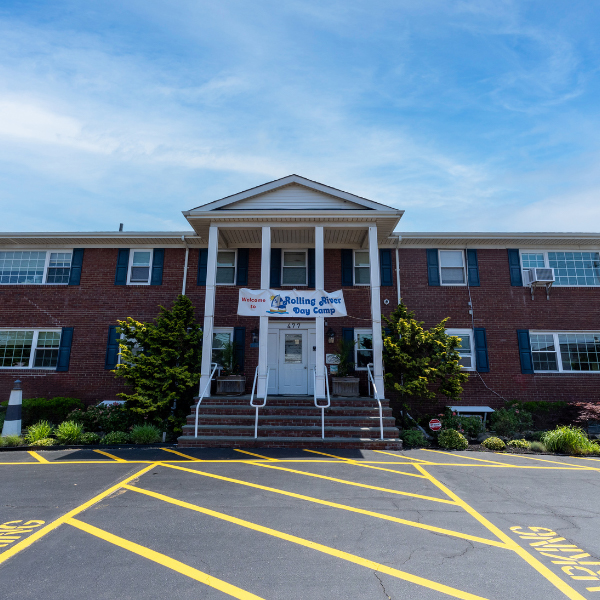 A two-story brick building with a sign reading "Rolling River Day Camp" above the entrance, flanked by shrubs and yellow-striped parking spaces in front.