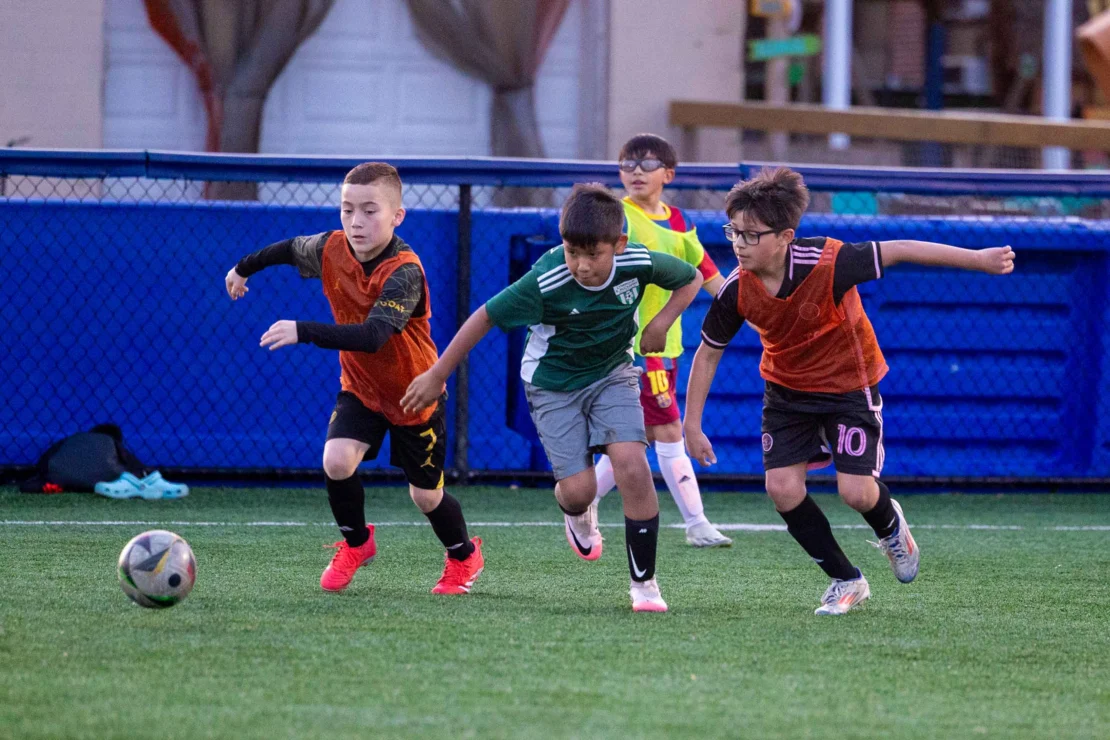 Three boys chase a soccer ball on an artificial turf field, while two other children watch from the background.