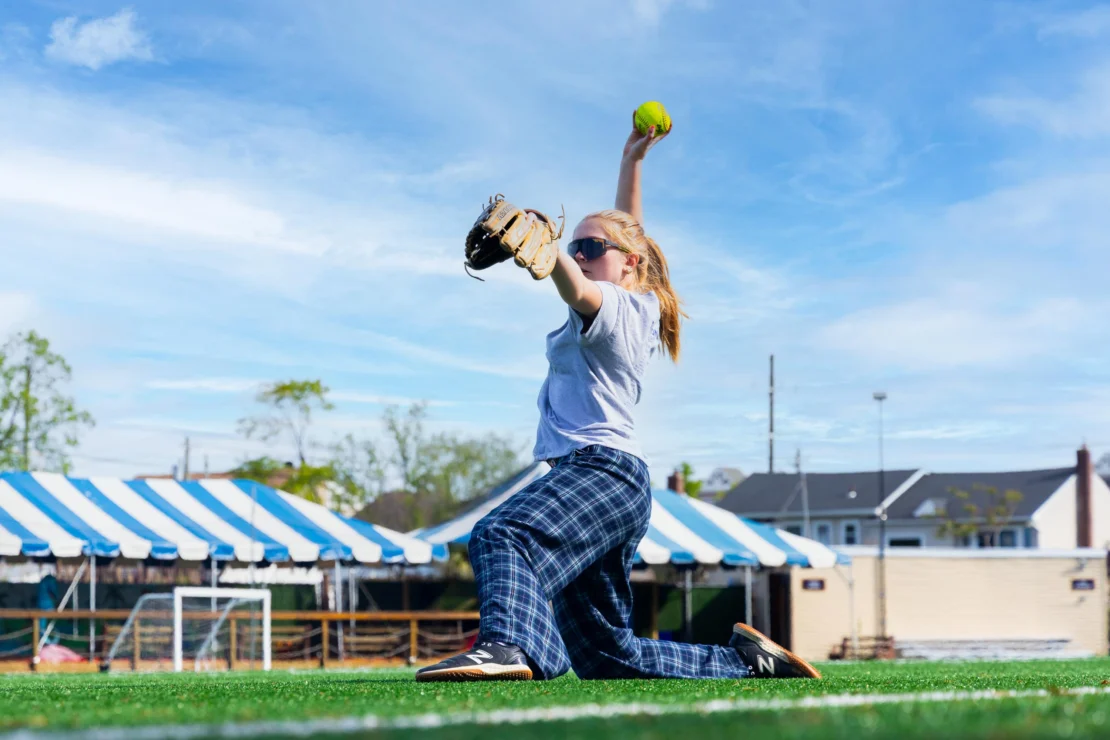 A person in plaid pants and sunglasses is kneeling on a sports field, preparing to throw a softball with one hand while wearing a glove on the other.