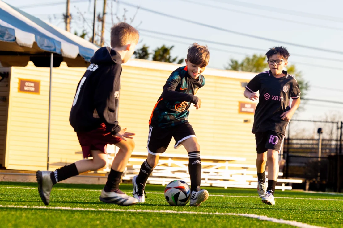 Three boys play soccer on a field, with one dribbling the ball while the other two approach him. A building and benches are in the background.