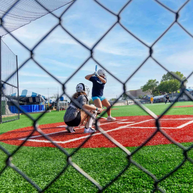 A youth baseball player prepares to bat while a catcher and umpire crouch behind home plate on a fenced-in artificial turf field.
