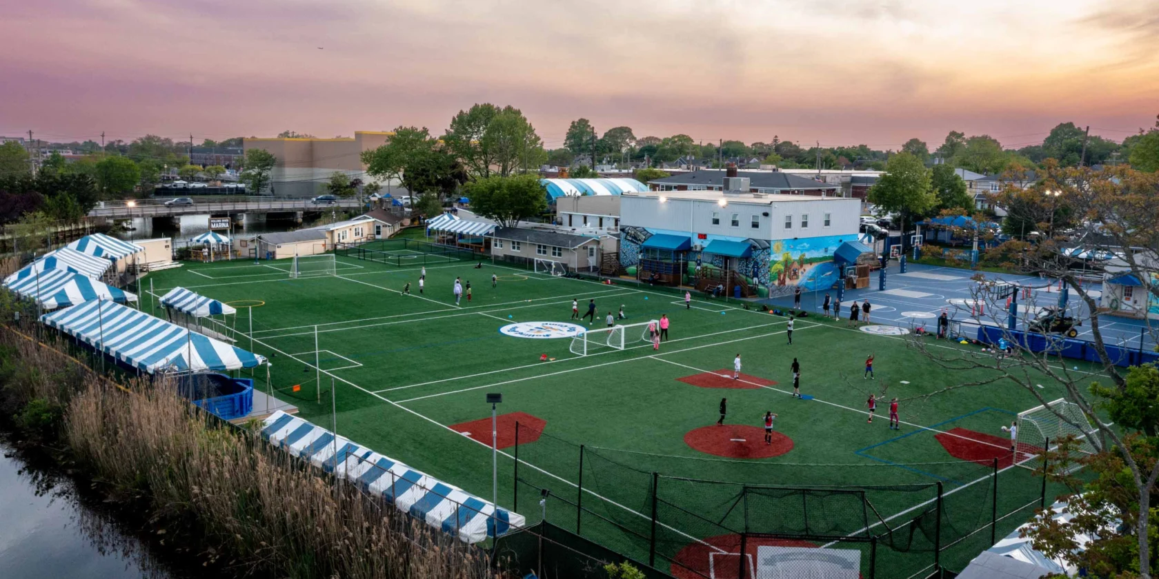 A sports complex with soccer fields and baseball areas, surrounded by striped tents and buildings, with people playing under a colorful sky at sunset.