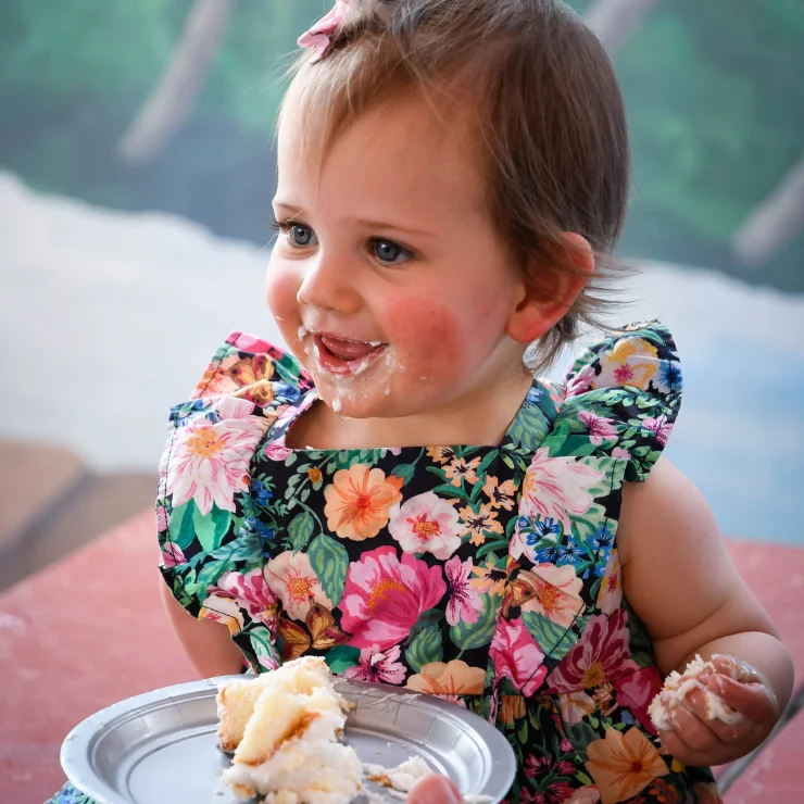 A toddler in a floral dress smiles with cake on her face and hands, holding a plate with a partially eaten piece of cake.