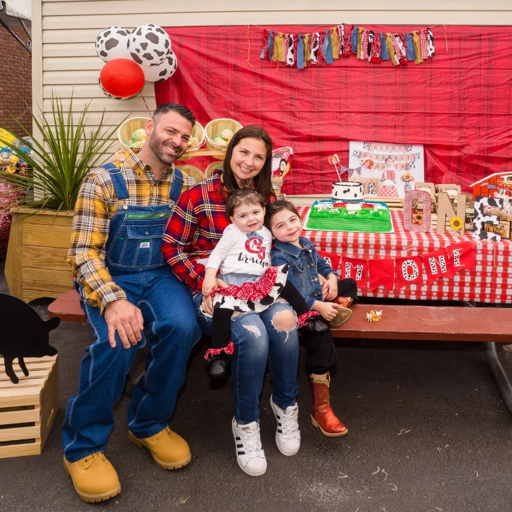 A family of four sits together at a farm-themed birthday party with red plaid decorations, a cake, and cow print accents.