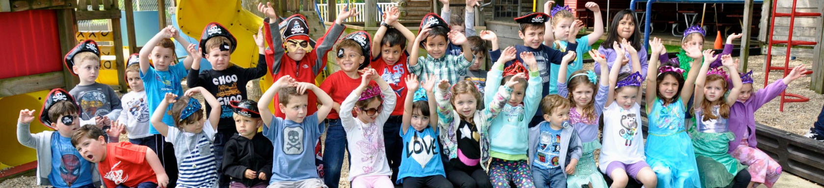 A large group of young children dressed in pirate hats and costumes sit and stand together outdoors, raising their hands and smiling for a group photo.
