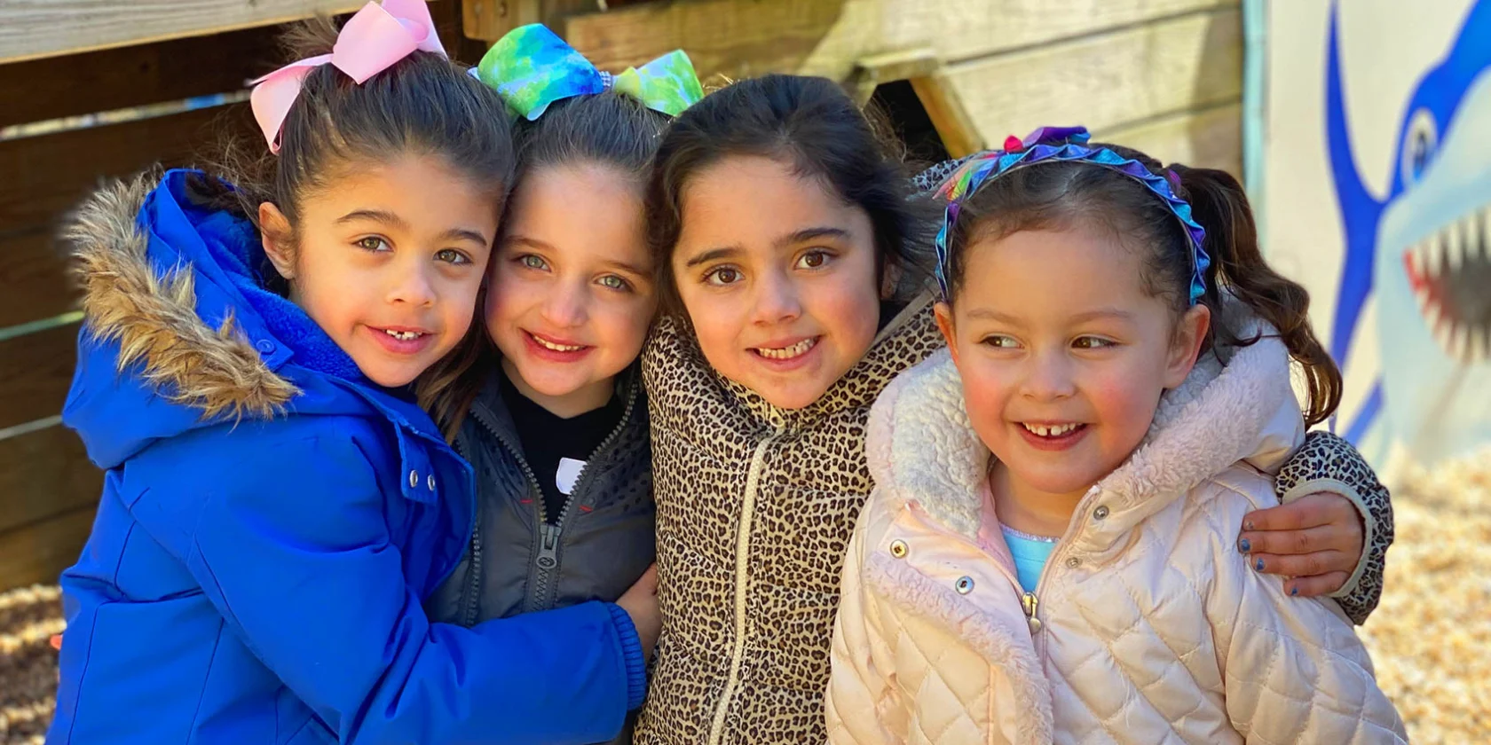 Four young girls in winter coats stand closely together, smiling outdoors near a wooden structure and a painted shark mural.