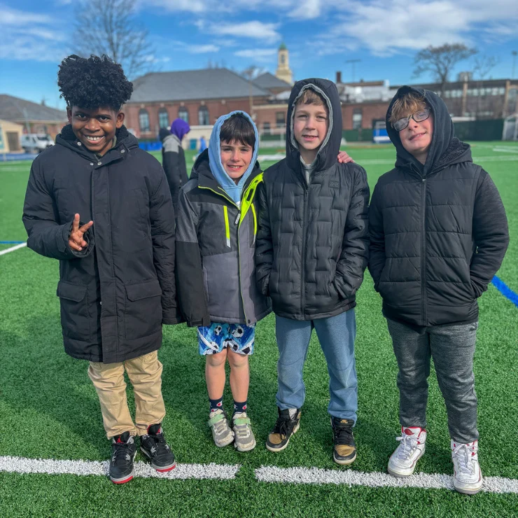 Four boys wearing jackets stand together on a green sports field, with buildings and a blue sky in the background.