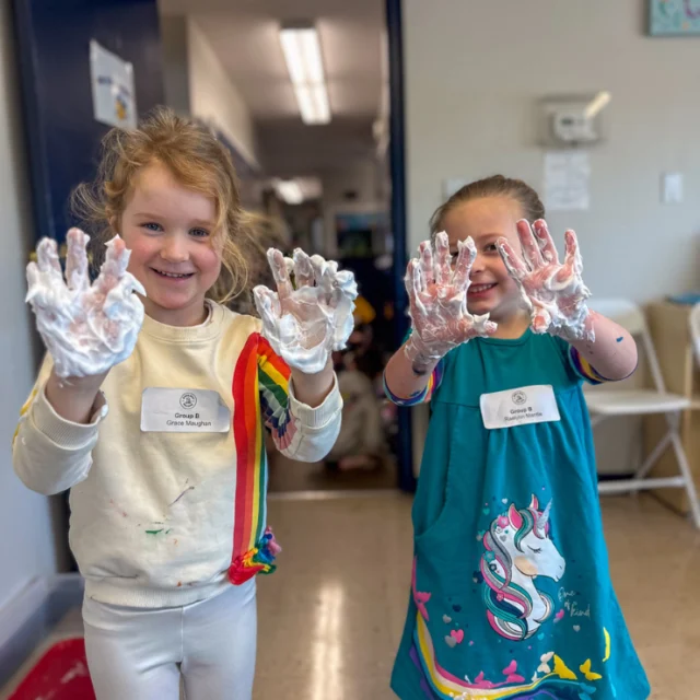 Two young children smile and hold up their hands covered in white foam, wearing nametags and colorful clothes, inside a classroom.