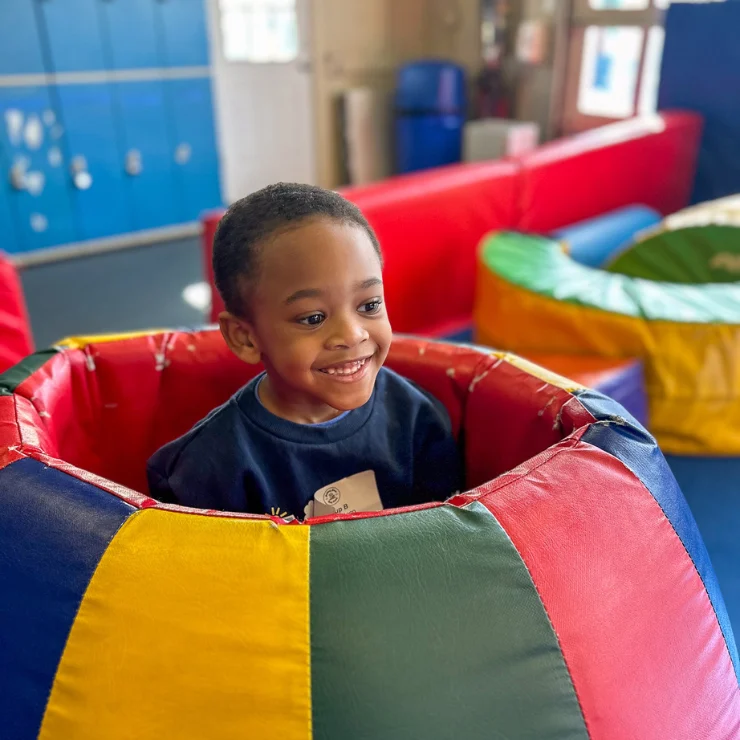 A young child smiles while sitting inside a colorful padded play tunnel in an indoor play area.