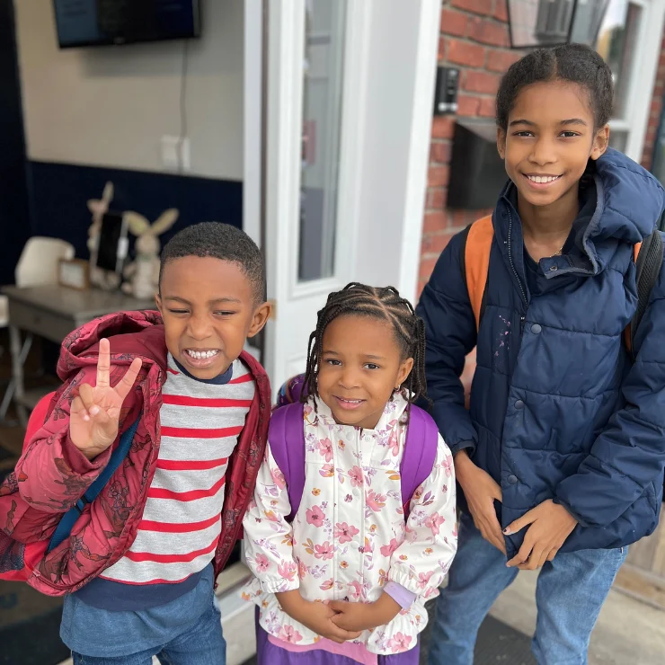 Three children wearing jackets and backpacks stand together outside a building. One child is making a peace sign and smiling, while the others look at the camera.