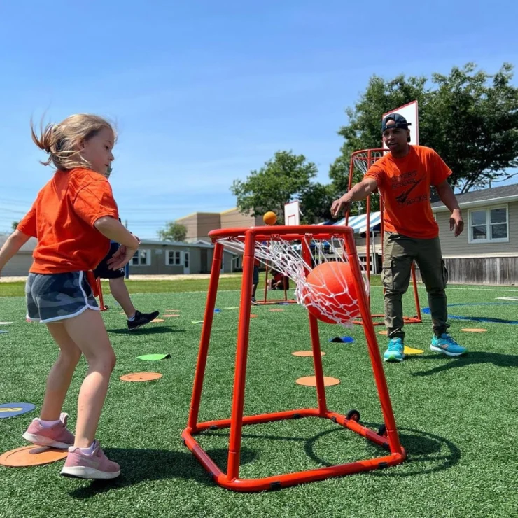 A young girl and an adult, both in orange shirts, play an outdoor basketball game on a turf field, with a ball going through a low basketball hoop.