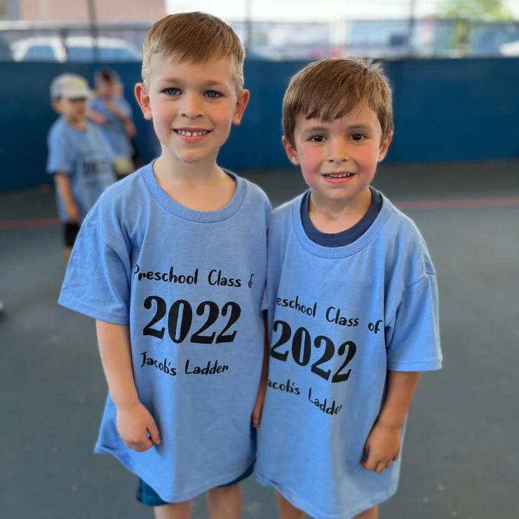 Two boys wearing matching “Preschool Class of 2022 Jacob’s Ladder” blue t-shirts stand side by side in a gym with other children in the background.