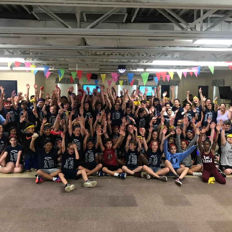 A large group of children and adults pose indoors with hands raised, wearing matching shirts and colorful headbands, under hanging bunting decorations.