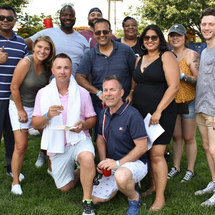 A group of people, some standing and some kneeling, pose and smile outdoors on grass during a sunny day. A few hold drinks or plates of food. Trees and event tents are visible in the background.