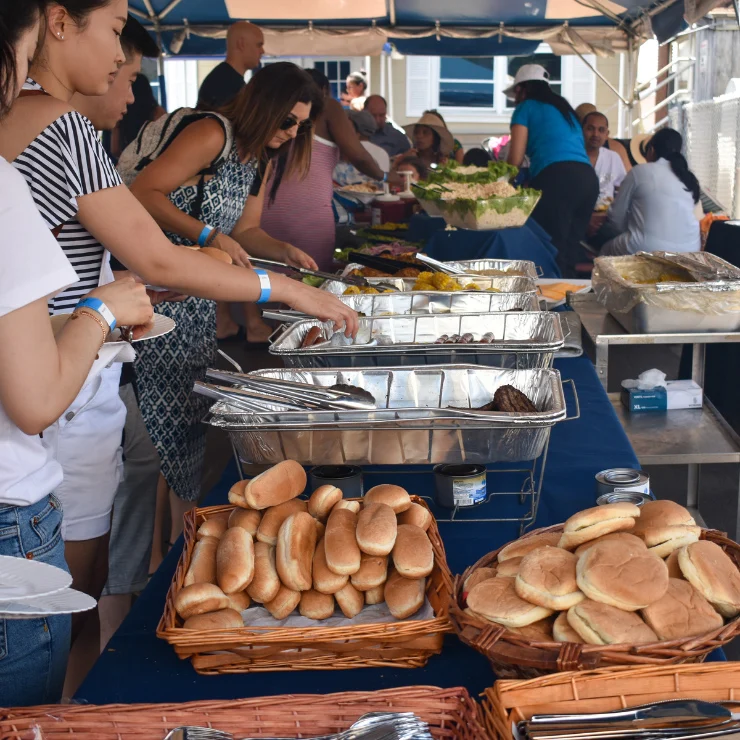 People serve themselves food from a buffet table outdoors, with trays of hot food, baskets of buns, and various dishes set out under a tent.