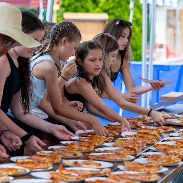 Several girls stand in line outdoors, each taking slices of pizza from a long table covered with plates of pizza.