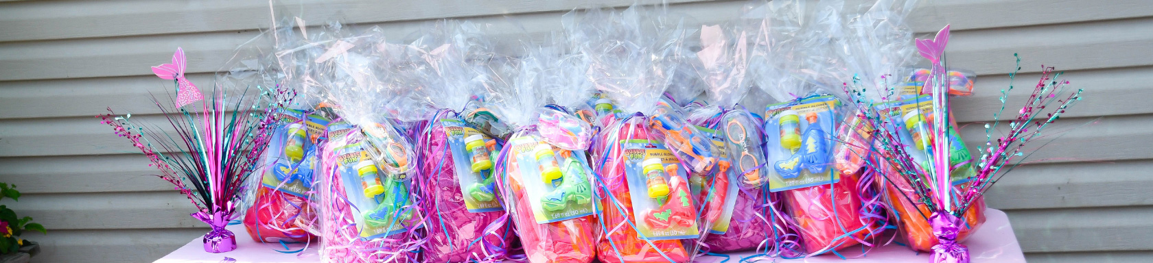 A row of colorful gift bags wrapped in clear plastic, each containing small toys and decorated with ribbons, displayed on a pink table in front of a gray siding wall.
