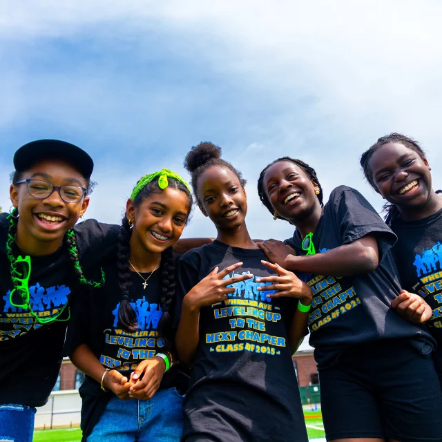 Five smiling girls wearing matching black shirts and neon green accessories pose outdoors on a grassy field under a partly cloudy sky.