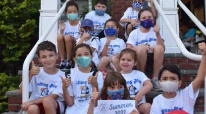 A group of children wearing masks and matching T-shirts sit on outdoor steps, some giving thumbs up, holding a “Summer 2021” sign.