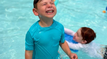 A young boy in a turquoise swim shirt stands smiling in a pool, holding a toy fish. Another child swims in the background.