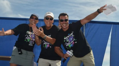 Three people wearing black event t-shirts and sunglasses smile and pose energetically in front of a blue curtain outdoors on a sunny day.