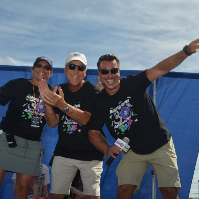 Three people wearing black event t-shirts and sunglasses smile and pose energetically in front of a blue curtain outdoors on a sunny day.