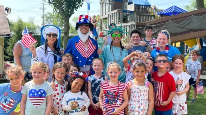 A group of adults and children in patriotic-themed outfits pose and smile outdoors at a playground, celebrating what appears to be a patriotic holiday.