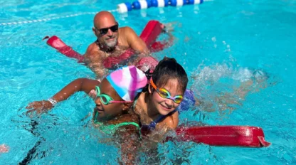 Two children wearing goggles and swim caps use kickboards in a pool, supervised by an adult man with sunglasses in the background.