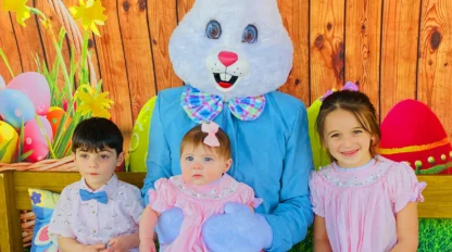 Three children pose with a person in an Easter Bunny costume in front of a wooden backdrop decorated with colorful eggs and flowers.