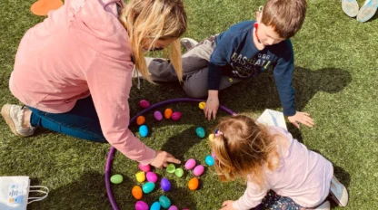 An adult and two children sit on grass sorting colorful plastic eggs inside a purple hoop.