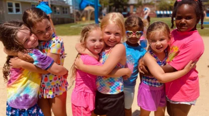 Seven young girls stand outdoors in a row, smiling and hugging each other, with a playground and volleyball net in the background on a sunny day.