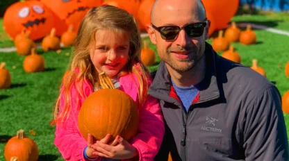 A man and a young girl holding a pumpkin pose outside among many pumpkins and large inflatable jack-o'-lanterns.