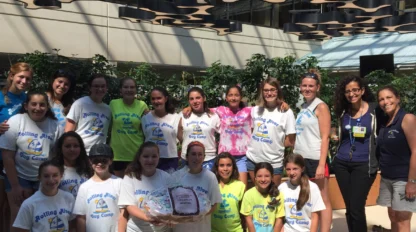 A group of girls and adults pose indoors, many wearing “Rolling River Day Camp” shirts. One girl holds a basket wrapped in cellophane. There are plants and modern ceiling decor in the background.