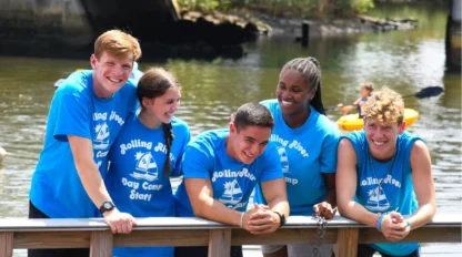 Five young adults in matching blue "Rolling River Day Camp Staff" shirts stand and lean on a wooden railing by the water, smiling and talking together on a sunny day.