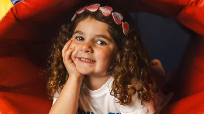 A young girl with curly hair and a heart-patterned headband smiles, leaning on her hand, inside a colorful play tunnel.